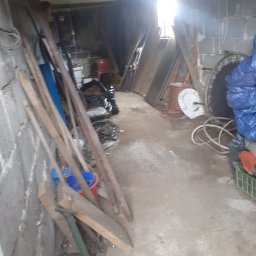 JUŻ w PORZĄDKU - Zagracone pomieszczenie gospodarcze z narzędziami, rurami i workami. Widoczne drewniane deski oparte o ścianę i nieporządek na podłodze.