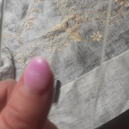 JUŻ w PORZĄDKU - Delikatny materiał z haftem kwiatowym w odcieniach szarości i beżu, częściowo zasłonięty przez palec z różowym lakierem i fragment narzędzia, prawdopodobnie podczas sprzątania.