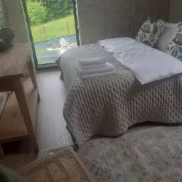 Jasne wnętrze sypialni z łóżkiem, stosem białych ręczników i widokiem na zieleń za oknem. Naturalne światło, drewniane elementy, stonowana kolorystyka.