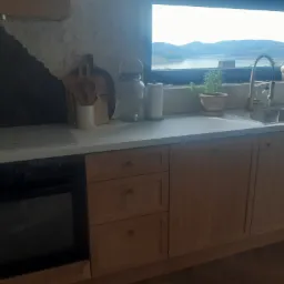 Jasna kuchnia z drewnianymi szafkami i widokiem na jezioro przez okno. Na blacie akcesoria kuchenne i zioła w doniczce, piekarnik w zabudowie, krzesło z czarnym oparciem.