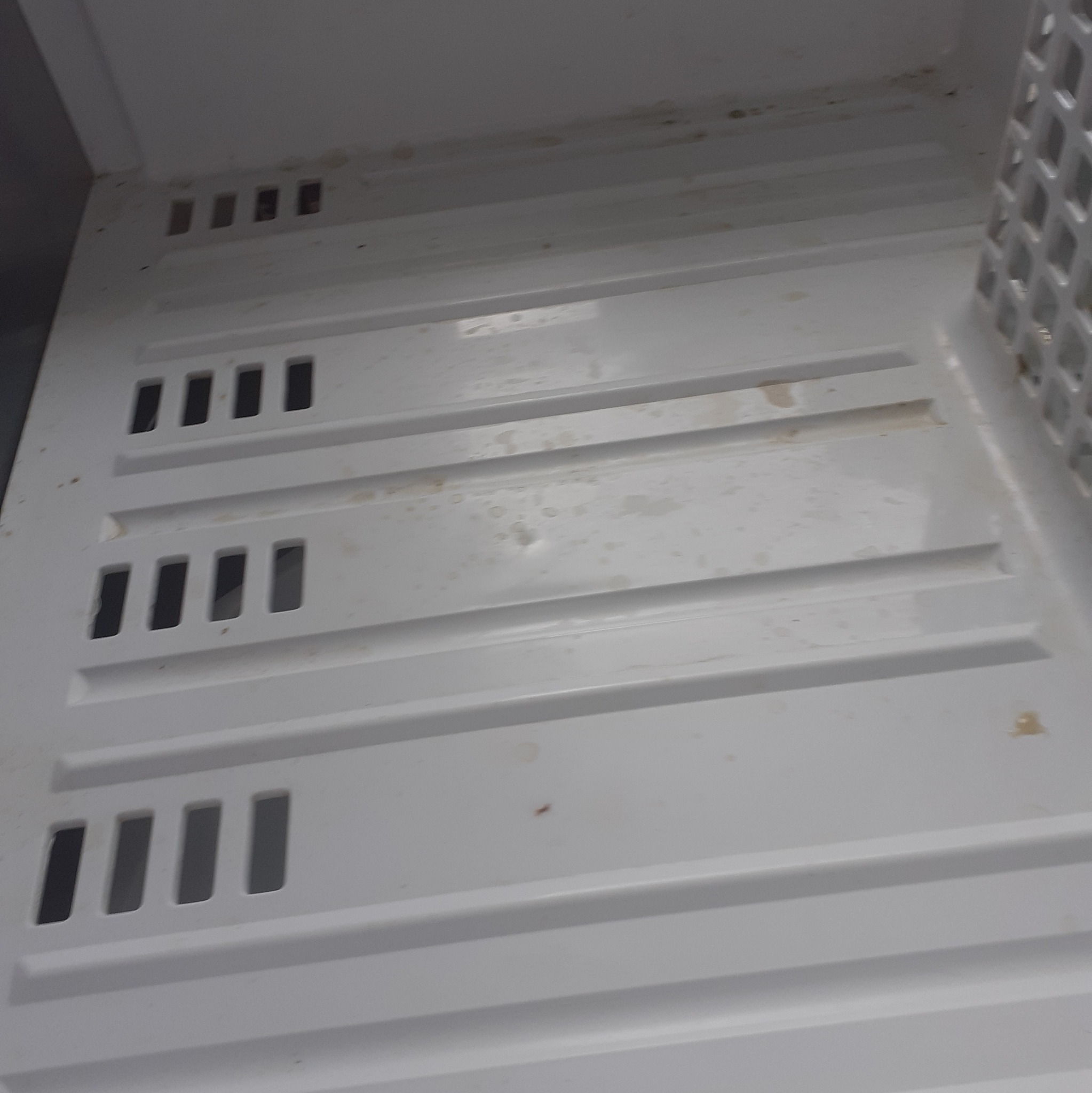 Brudna, biała półka w lodówce z licznymi plamami i zaciekami, widoczne otwory wentylacyjne. Zaniedbana higiena wnętrza chłodziarki.