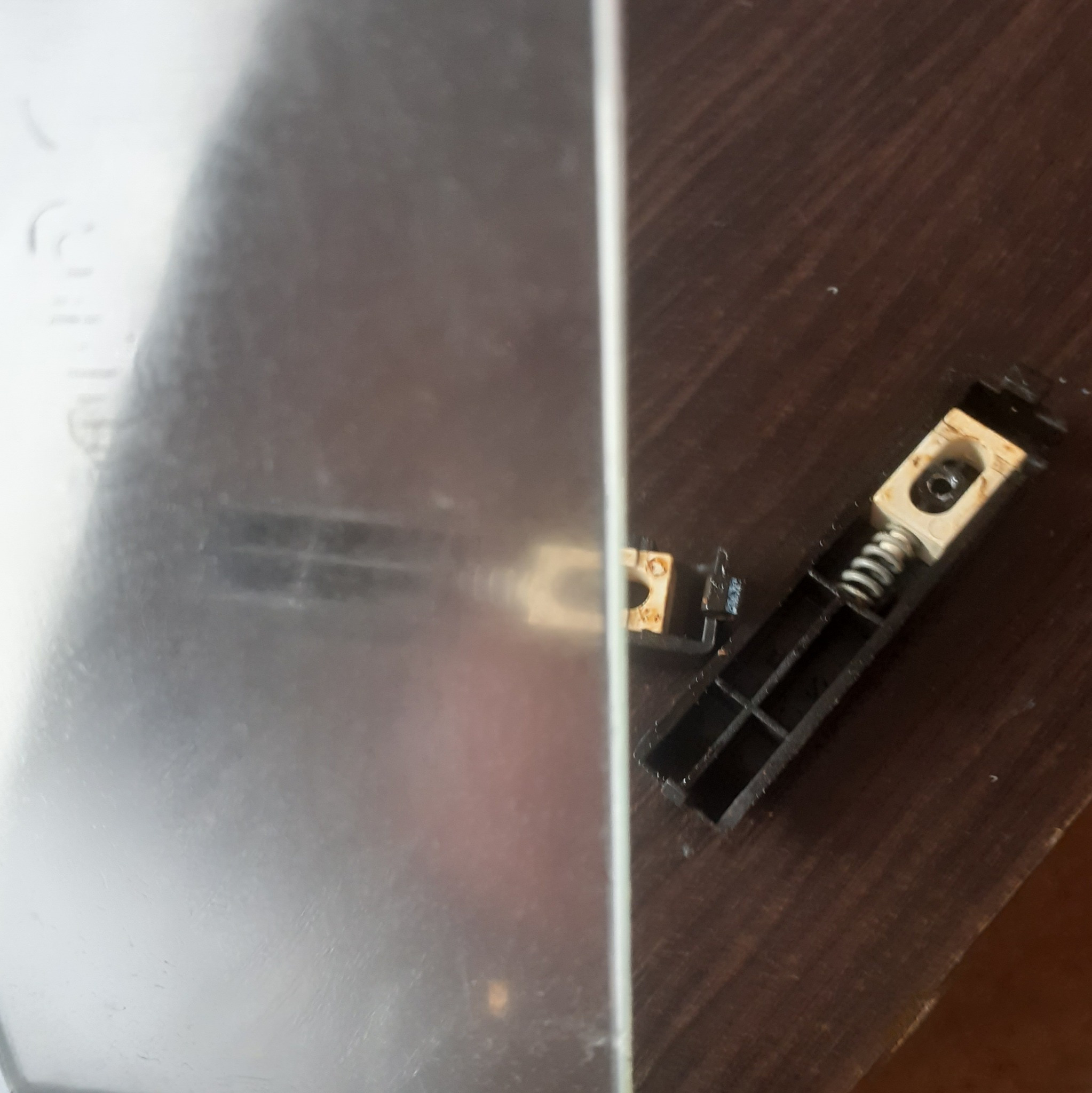 Uszkodzony element mechaniczny z metalową płytą, widoczny na drewnianej powierzchni. Sprężyna i plastikowa obudowa sugerują awarię urządzenia.