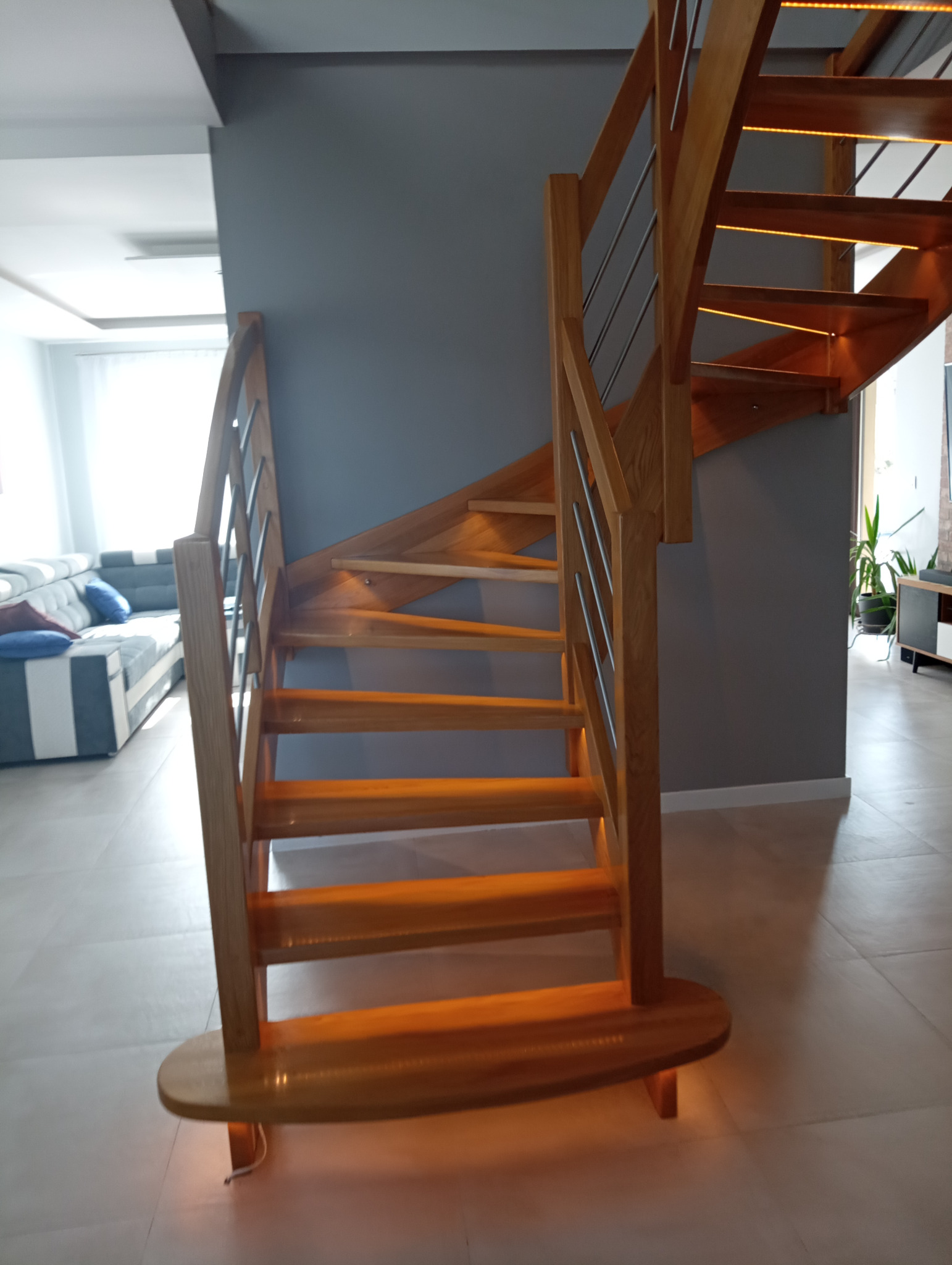 Drewniane schody kręcone z podświetleniem LED, metalowe barierki, szare ściany i fragment salonu z sofą w tle. Nowoczesny design.
