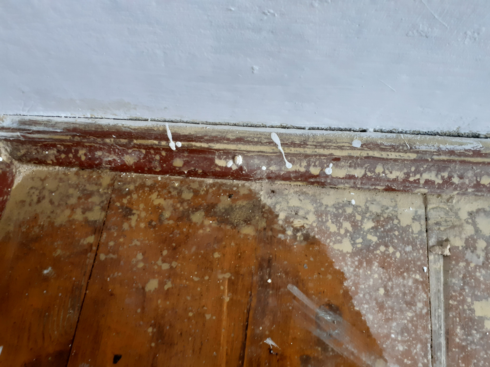 Zabrudzona farbą podłoga z brązowych desek i biała ściana po malowaniu, widoczne krople i zacieki farby. Niewyczyszczone pozostałości po remoncie.