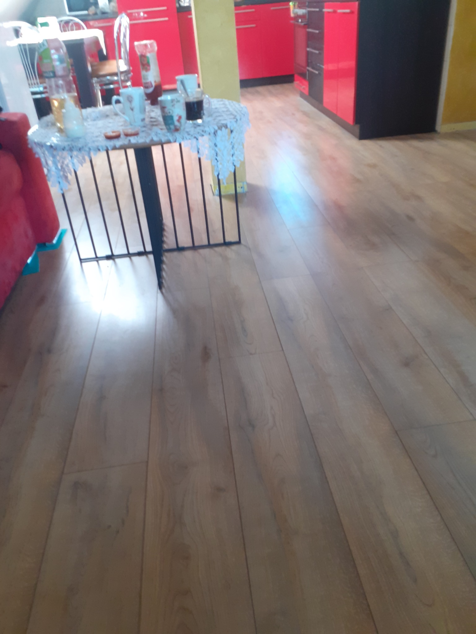 Błyszcząca, drewniana podłoga w salonie z czerwonymi akcentami kuchennymi w tle, stół z koronkową serwetą i naczyniami.