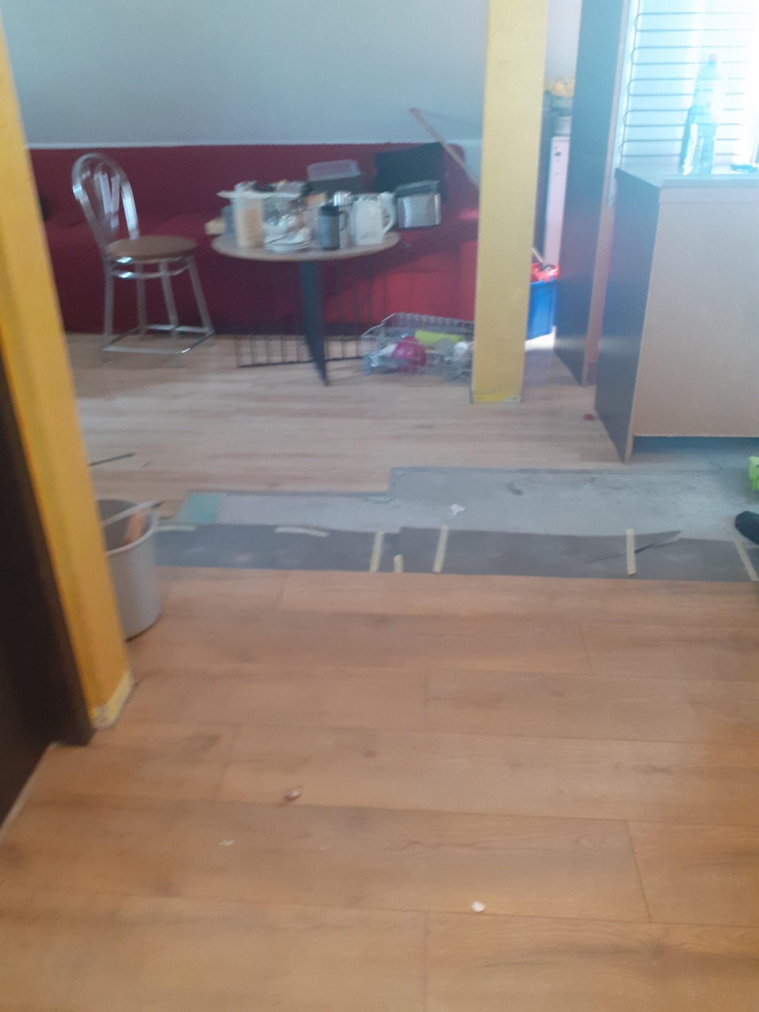Wnętrze pokoju z drewnianą podłogą w trakcie prac. Widoczne przygotowania do położenia podłogi, fragmenty podkładu i resztki po remoncie. Na stole naczynia i przedmioty.
