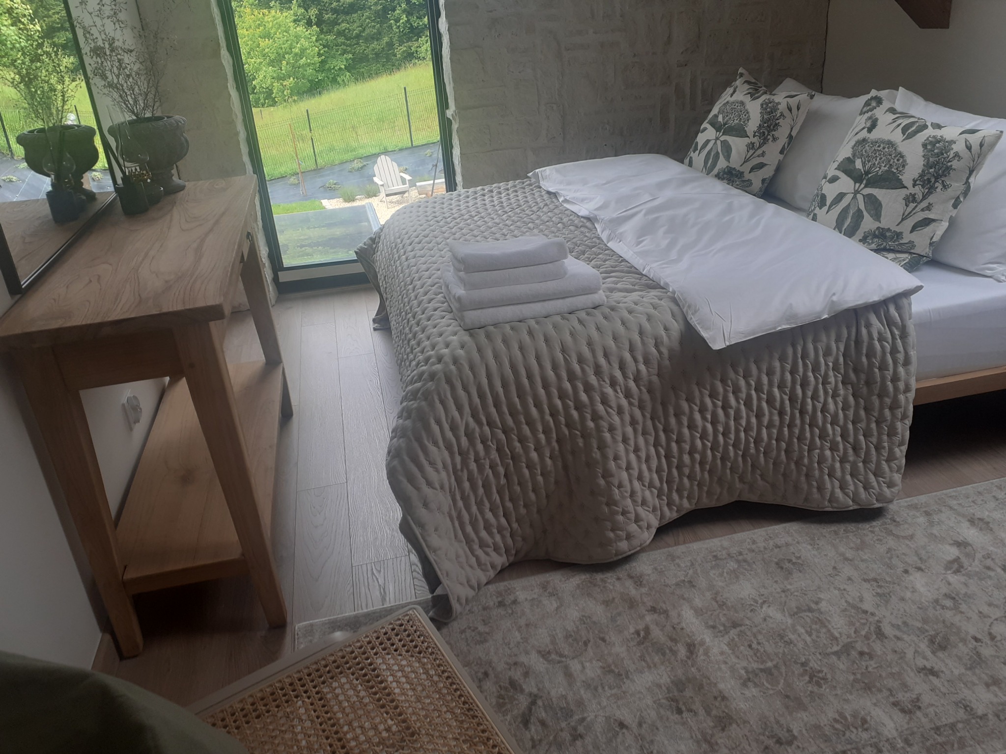 Jasne wnętrze sypialni z łóżkiem, stosem białych ręczników i widokiem na zieleń za oknem. Naturalne światło, drewniane elementy, stonowana kolorystyka.