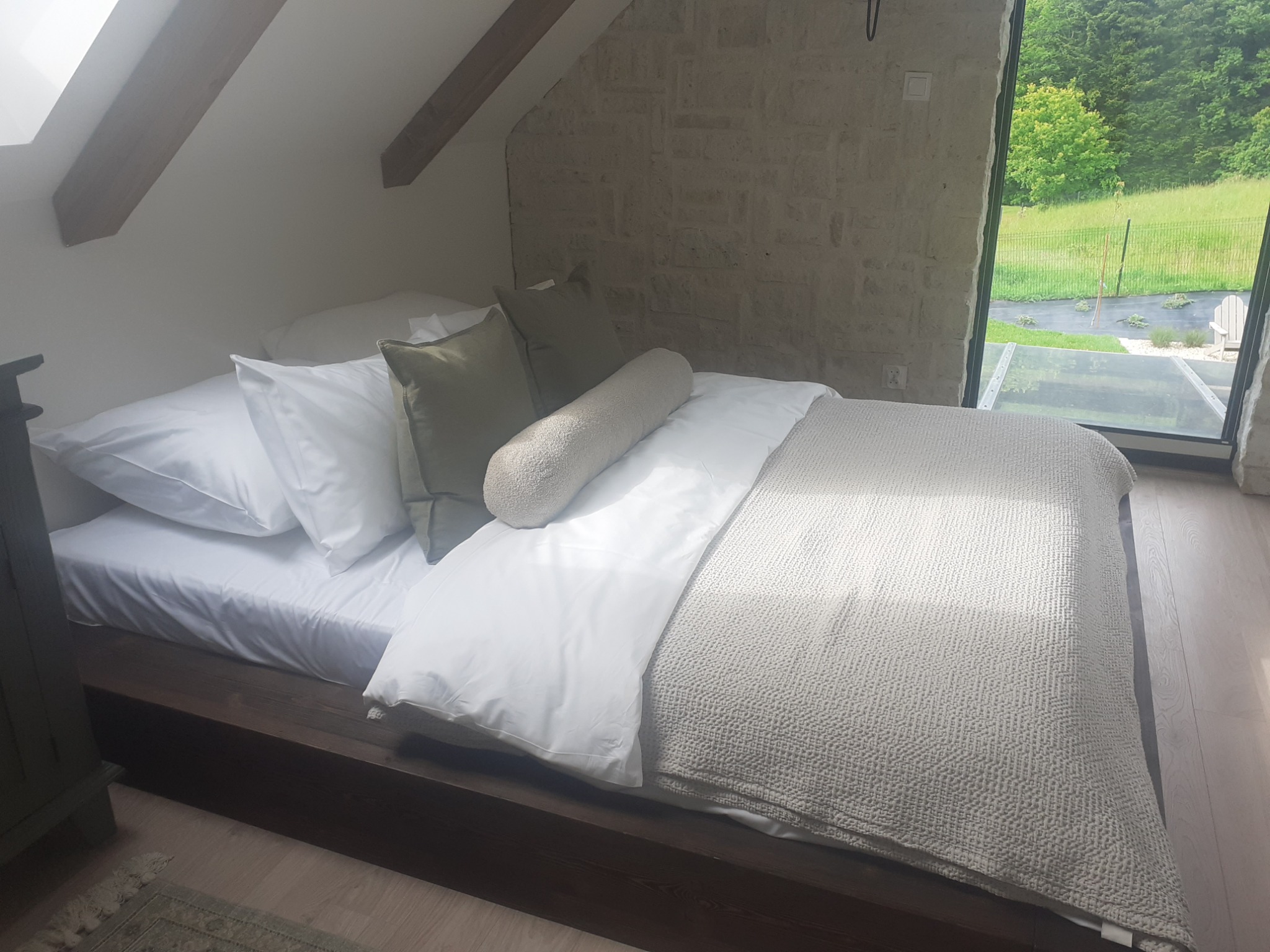 Jasna sypialnia na poddaszu z widokiem na zieleń. Kamienna ściana i duże okno w tle. Minimalistyczne łóżko z białą pościelą i dekoracyjnymi poduszkami.