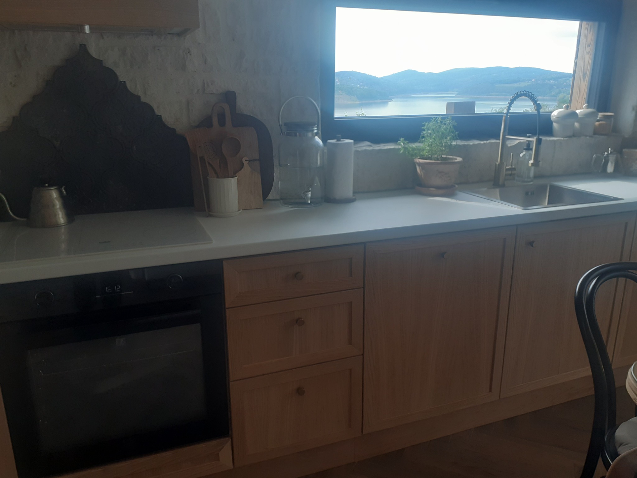 Jasna kuchnia z drewnianymi szafkami i widokiem na jezioro przez okno. Na blacie akcesoria kuchenne i zioła w doniczce, piekarnik w zabudowie, krzesło z czarnym oparciem.