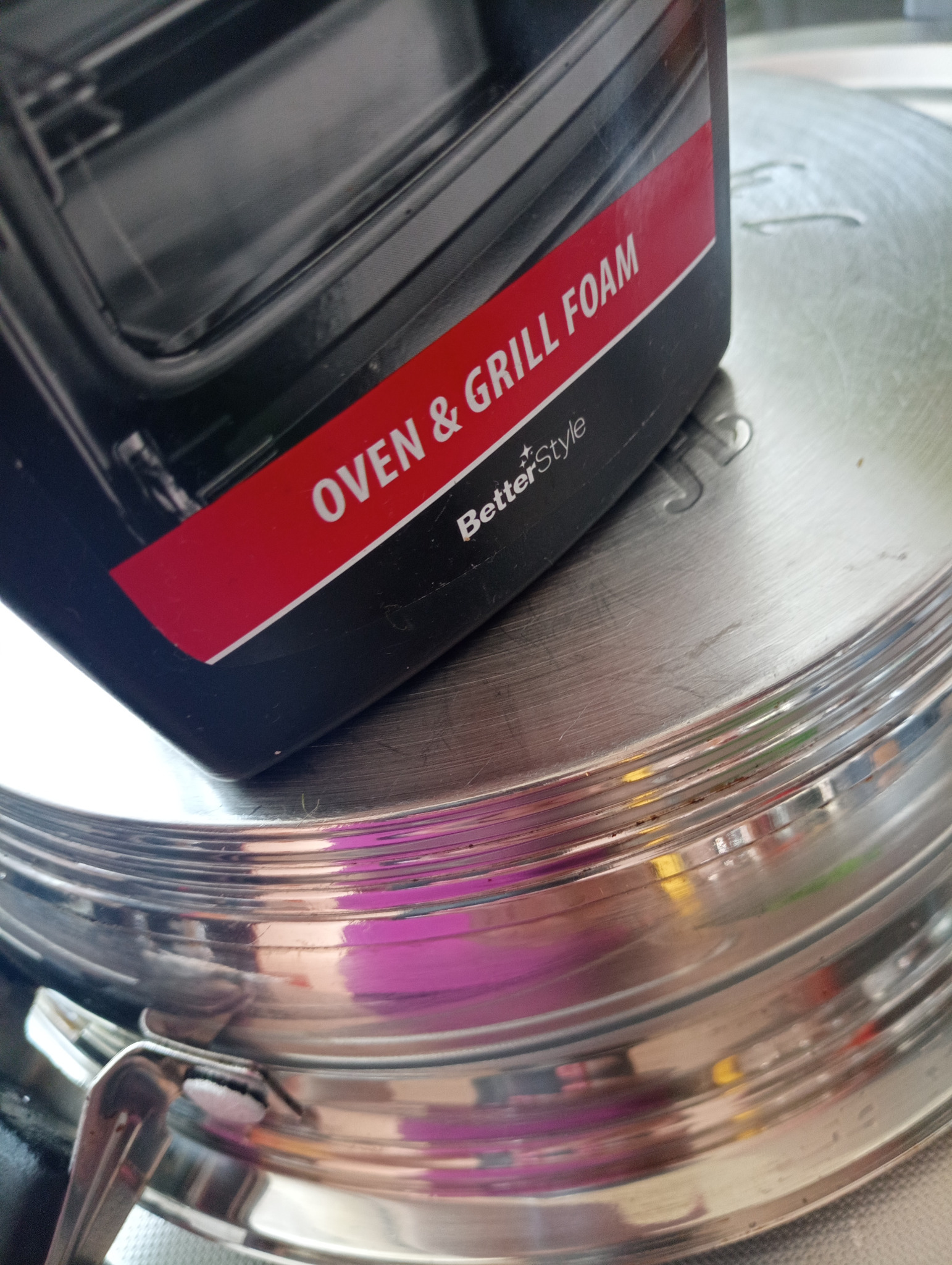 Płyn do czyszczenia piekarników i grilli BetterStyle na metalowej pokrywie. Produkt do sprzątania, czarna butelka z czerwonym paskiem i białym napisem.