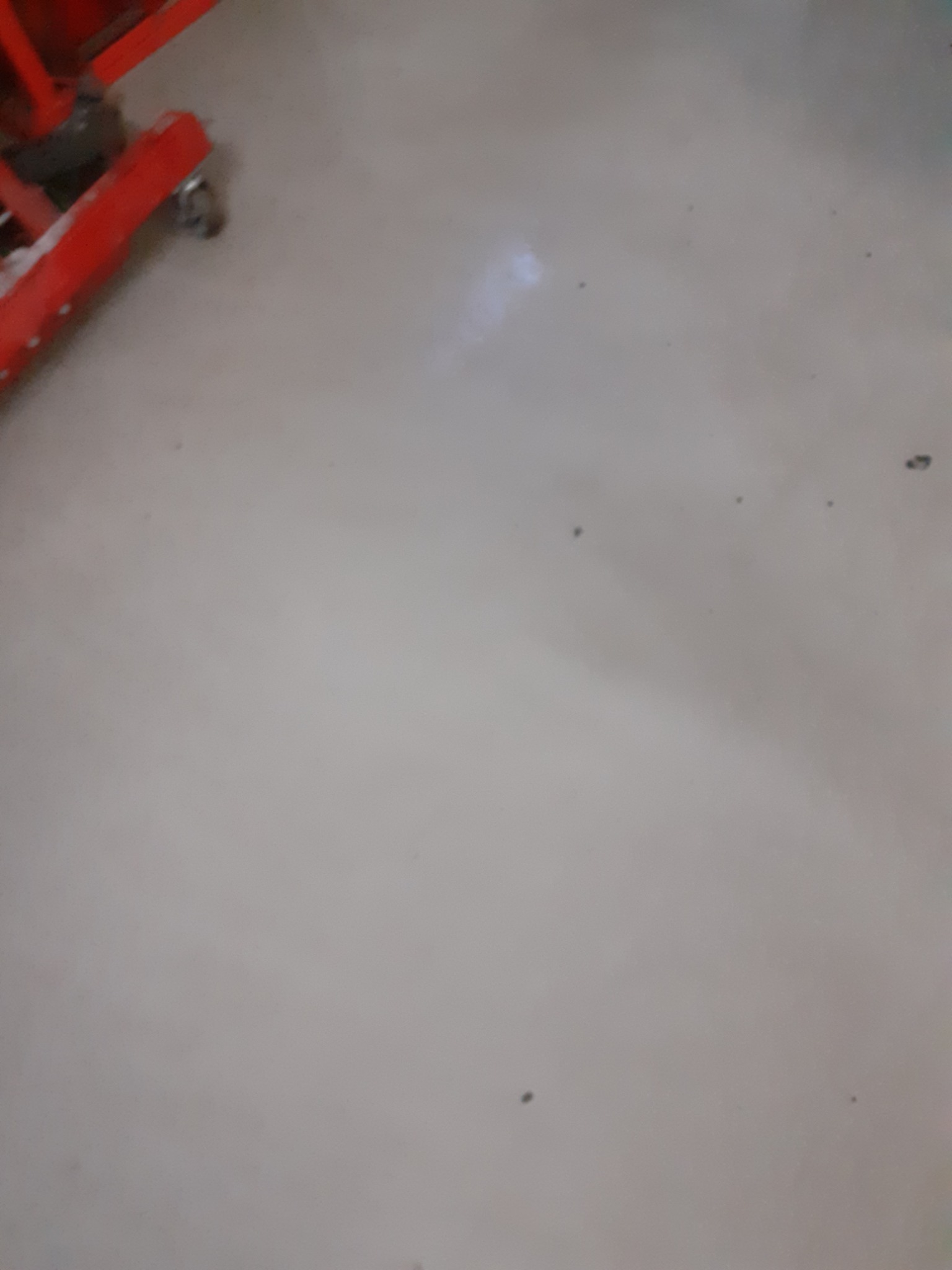 Jasna, gładka podłoga po sprzątaniu z fragmentem czerwonego wózka w lewym górnym rogu. Minimalistyczne ujęcie, skupione na czystości powierzchni.