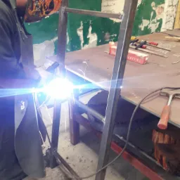 Spawanie metalowej konstrukcji ramowej z użyciem maski spawalniczej z motywem płomieni, widoczny stół roboczy z narzędziami i niedokończona ściana w tle.