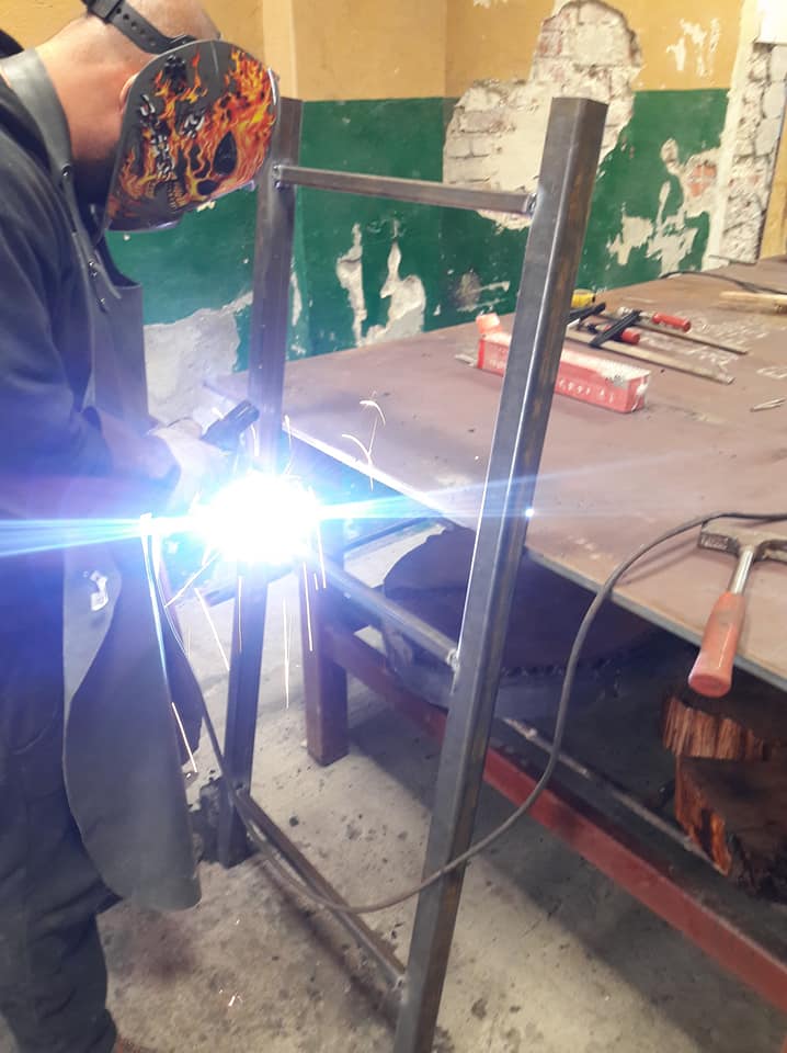 Spawanie metalowej konstrukcji ramowej z użyciem maski spawalniczej z motywem płomieni, widoczny stół roboczy z narzędziami i niedokończona ściana w tle.