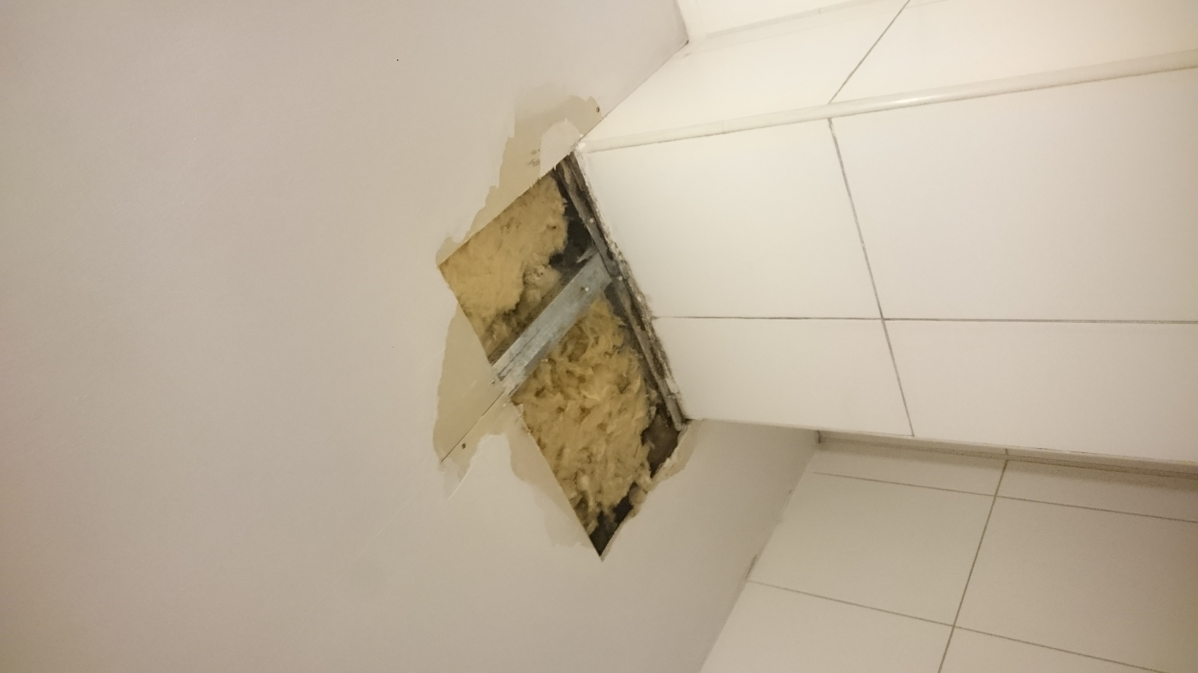 Otwór w suficie odsłaniający izolację z wełny mineralnej i metalową konstrukcję wsporczą, widoczne białe płytki na ścianie i fragment sufitu przed naprawą