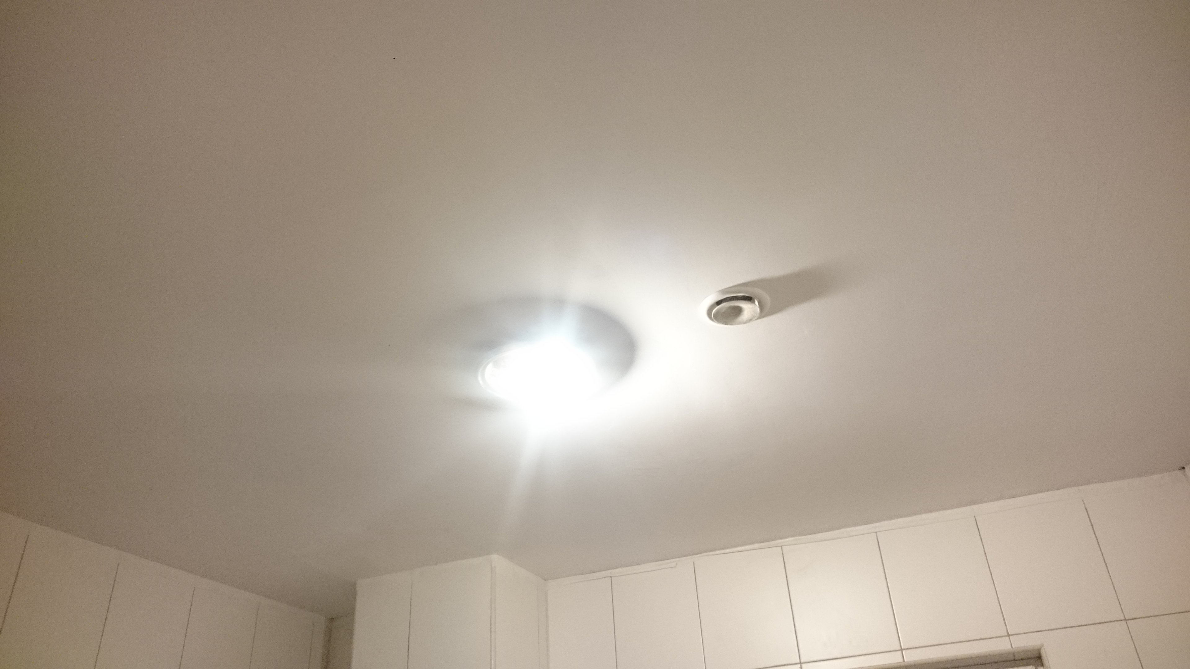 Biały sufit z zamontowanym oświetleniem punktowym i kratką wentylacyjną, widoczne także fragmenty białych szafek kuchennych.