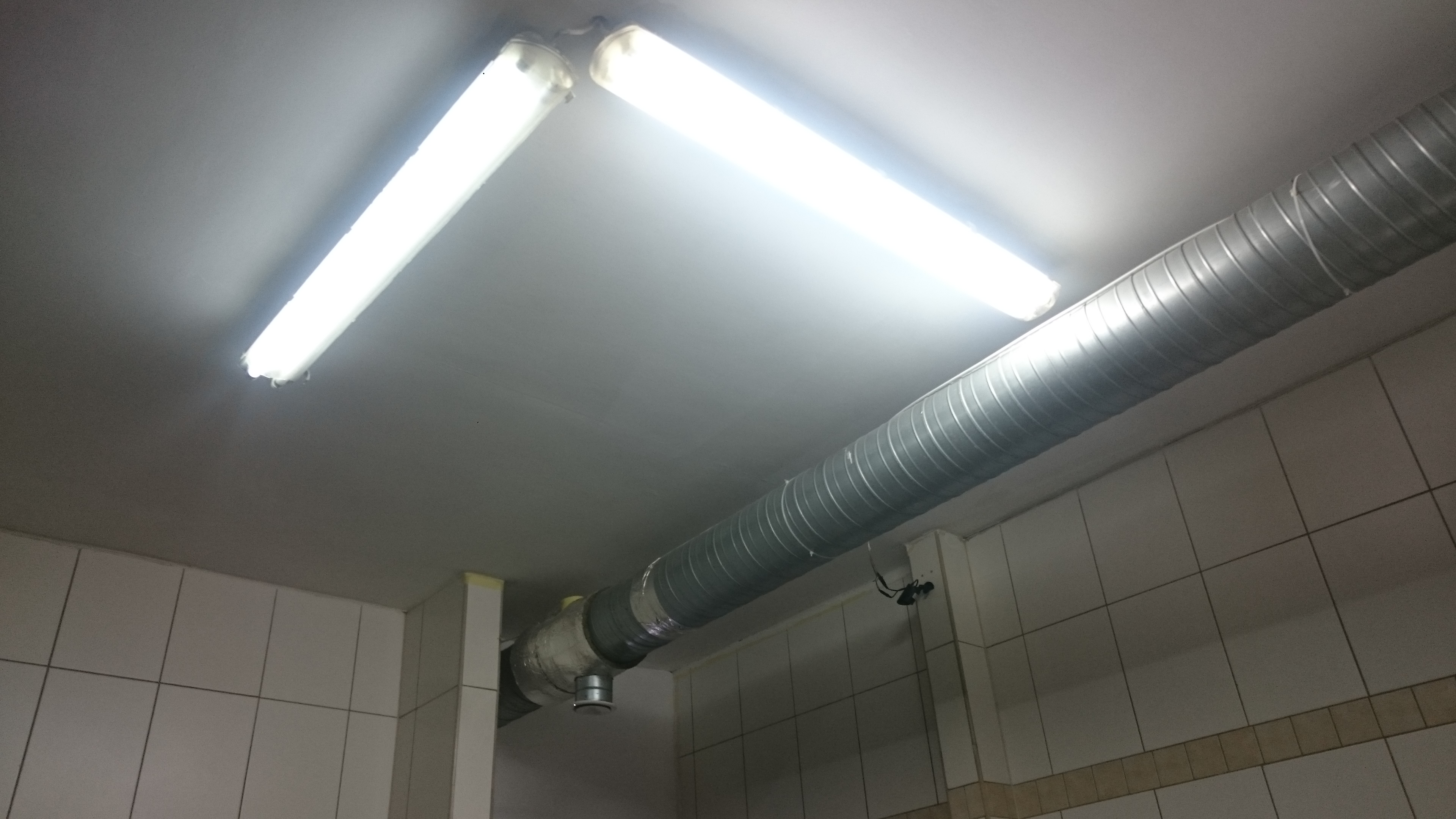 Oświetlenie LED i metalowy przewód wentylacyjny pod sufitem w pomieszczeniu wyłożonym białymi kafelkami.