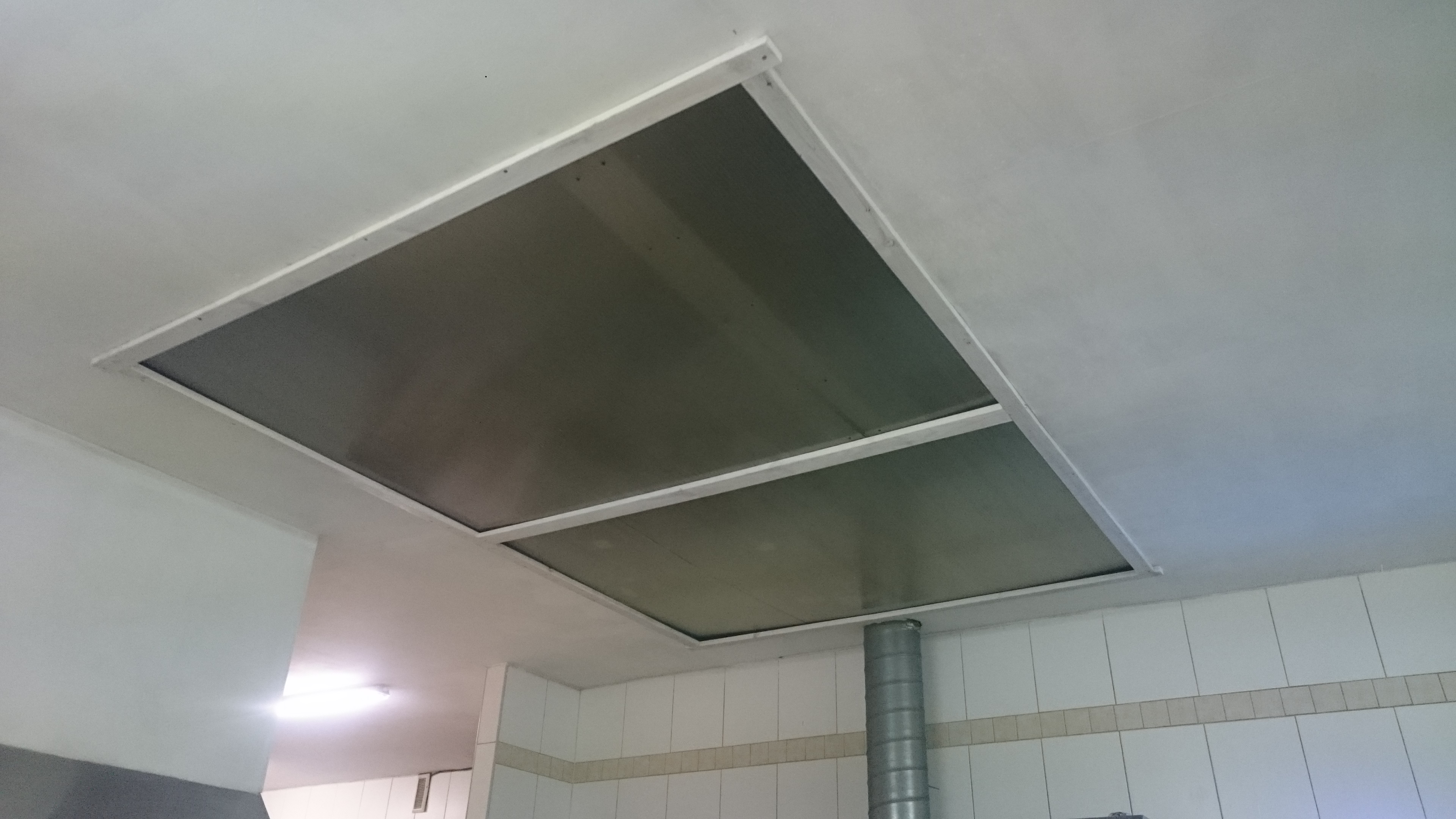 Dwa kwadratowe panele sufitowe zamontowane w białej ramie na suficie, widoczne rury wentylacyjne i białe płytki na ścianie w tle.