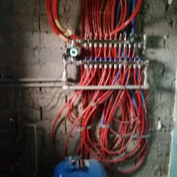 Rozdzielacz ogrzewania podłogowego z czerwonymi i niebieskimi rurami zamontowany na surowej ścianie z cegieł, obok fragmentu ściany pokrytej płytą gipsowo-kartonową.