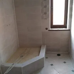 Wnętrze łazienki w trakcie prac wykończeniowych: kabina prysznicowa z niskim brodzikiem, jasne płytki na ścianach i podłodze z dekoracyjnymi wstawkami, okno z drewnianą ramą, widoczne narzędzia...