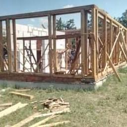 Pan- Bud - Wznoszenie konstrukcji szkieletowej budynku z drewnianych belek na fundamencie, widoczne fragmenty muru z cegły i pustaków, na trawie leżą resztki drewna i żółta izolacja.