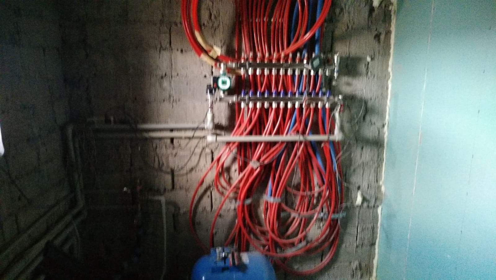 Rozdzielacz ogrzewania podłogowego z czerwonymi i niebieskimi rurami zamontowany na surowej ścianie z cegieł, obok fragmentu ściany pokrytej płytą gipsowo-kartonową.