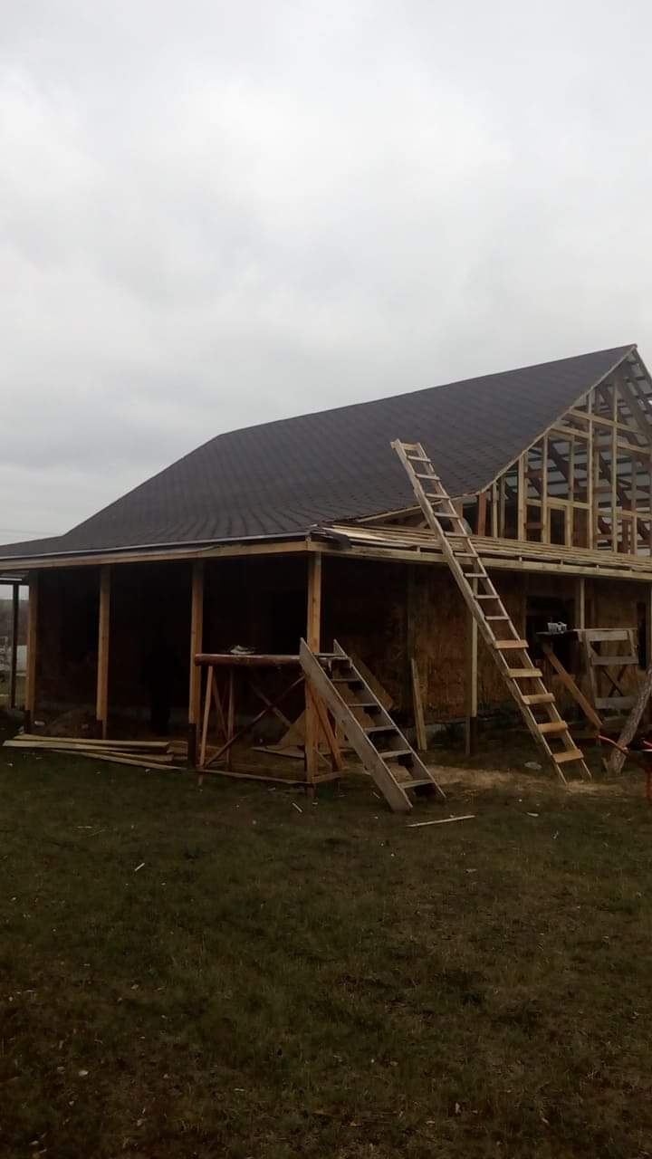 Wznoszenie drewnianej konstrukcji domu jednorodzinnego z częściowo ukończonym dachem, widoczne rusztowania i drabiny na tle pochmurnego nieba.