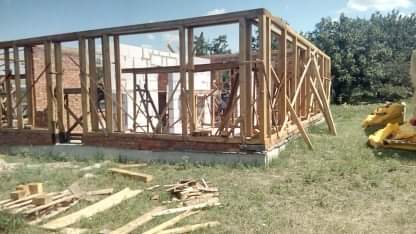 Wznoszenie konstrukcji szkieletowej budynku z drewnianych belek na fundamencie, widoczne fragmenty muru z cegły i pustaków, na trawie leżą resztki drewna i żółta izolacja.