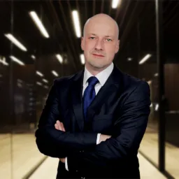 Portret mężczyzny w ciemnym garniturze i niebieskim krawacie, stojącego na tle rozmytego, nowoczesnego biura z podświetlanymi elementami.