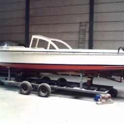 Biała łódź motorowa z czerwonym dnem, umieszczona na szarej przyczepie wewnątrz hali z betonowymi ścianami. Widoczna drabina w tle.