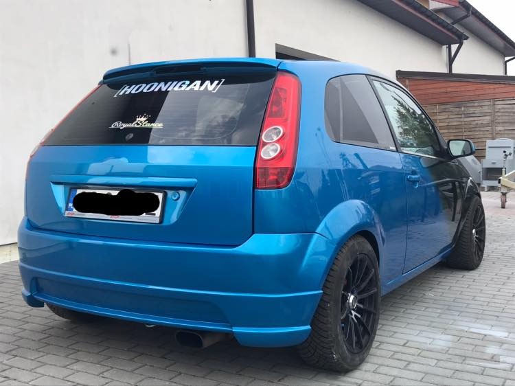 Niebieski Ford Fiesta po tuningu, widok z tyłu, z naklejkami 'Hoonigan' i 'Royal Stance' na tylnej szybie, czarne felgi, sportowy wydech.