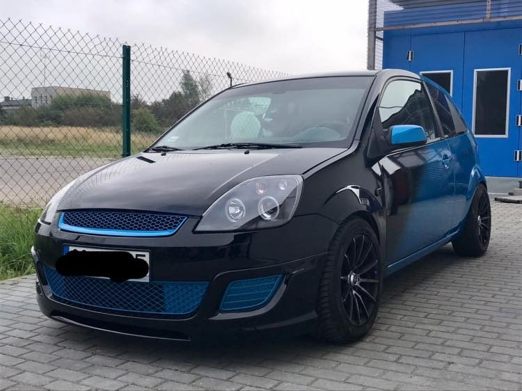 Czarno-niebieski Ford Fiesta po tuningu, z niebieskimi akcentami na grillu, lusterkach i tylnej części nadwozia, zaparkowany na kostce brukowej przed niebieskim budynkiem.
