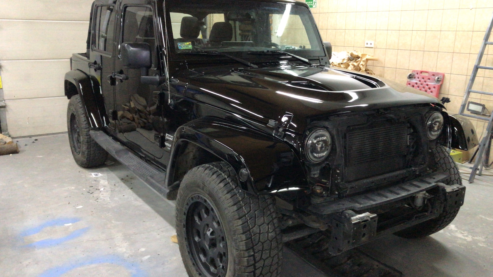 Czarny Jeep Wrangler z częściowo zdemontowanym przodem, widoczne nowe reflektory i błyszcząca karoseria po lakierowaniu, w tle garaż.