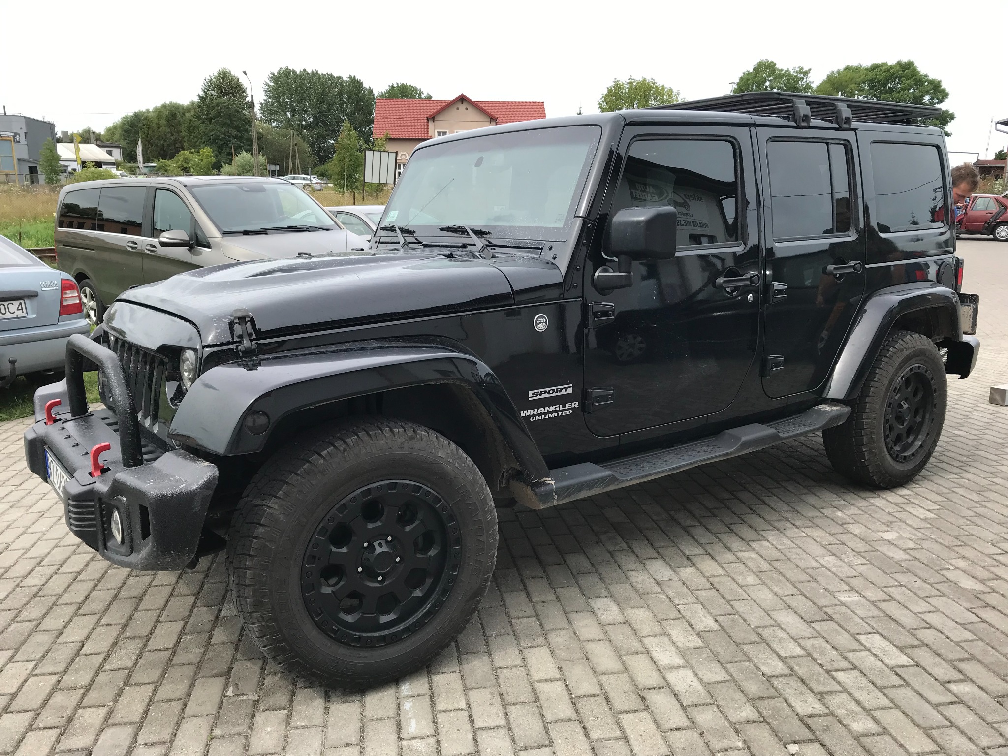 Czarny Jeep Wrangler Unlimited z bagażnikiem dachowym i orurowaniem przedniego zderzaka, zaparkowany na kostce brukowej.