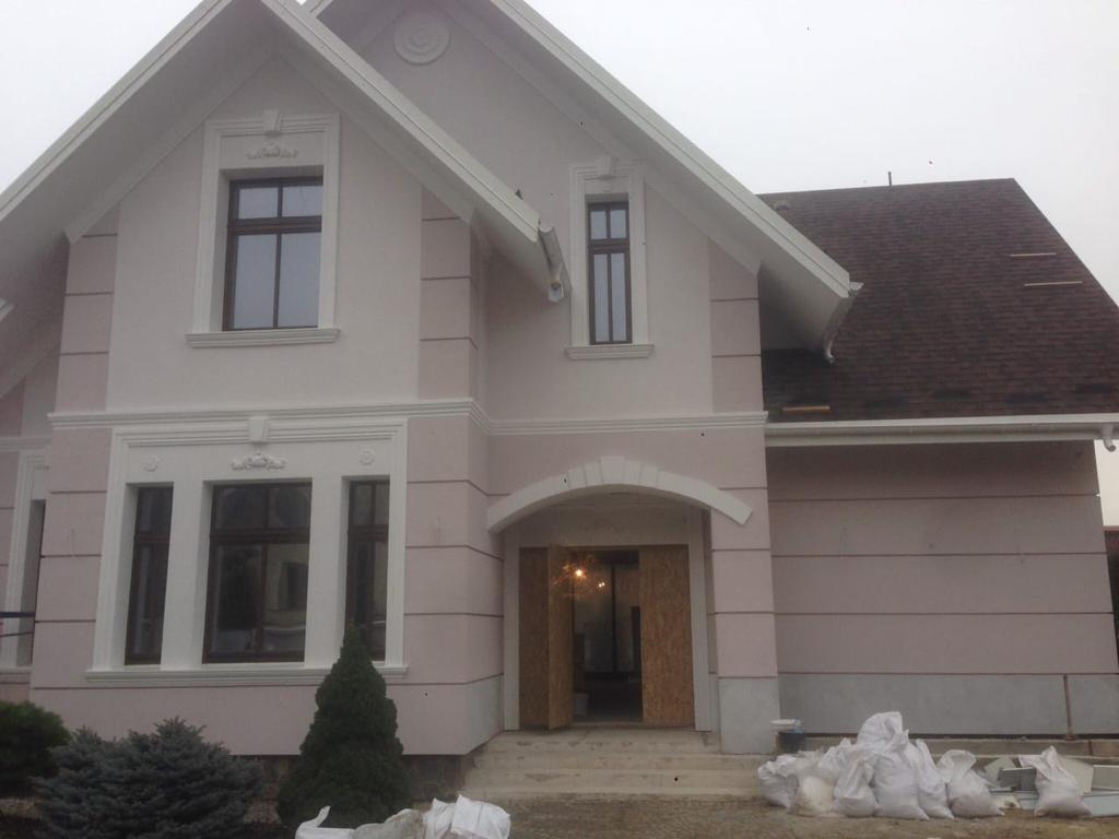 Dwupiętrowy dom z jasnoróżową elewacją i białymi zdobieniami wokół okien i dachu, brązowy dach z bitumicznej dachówki, worki z materiałem budowlanym przed wejściem.