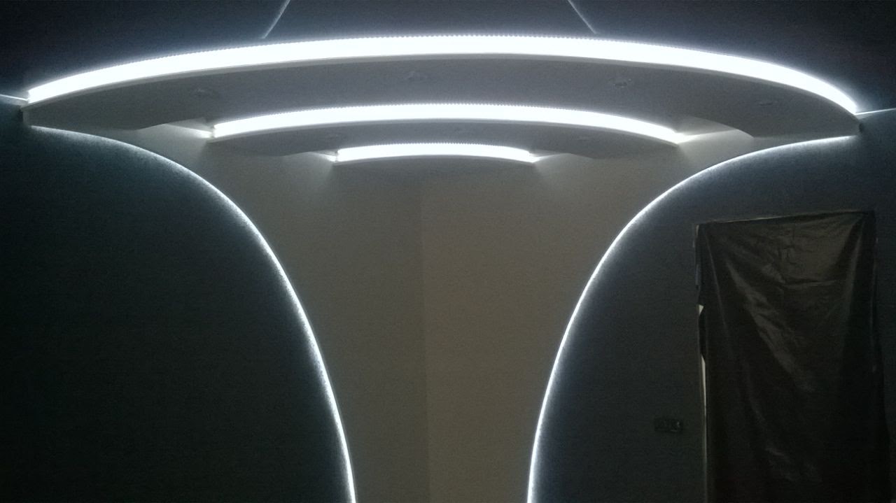 Półokrągłe wnęki w suficie z oświetleniem LED, widoczna ściana w rogu pomieszczenia i zasłonięte czarną folią drzwi.