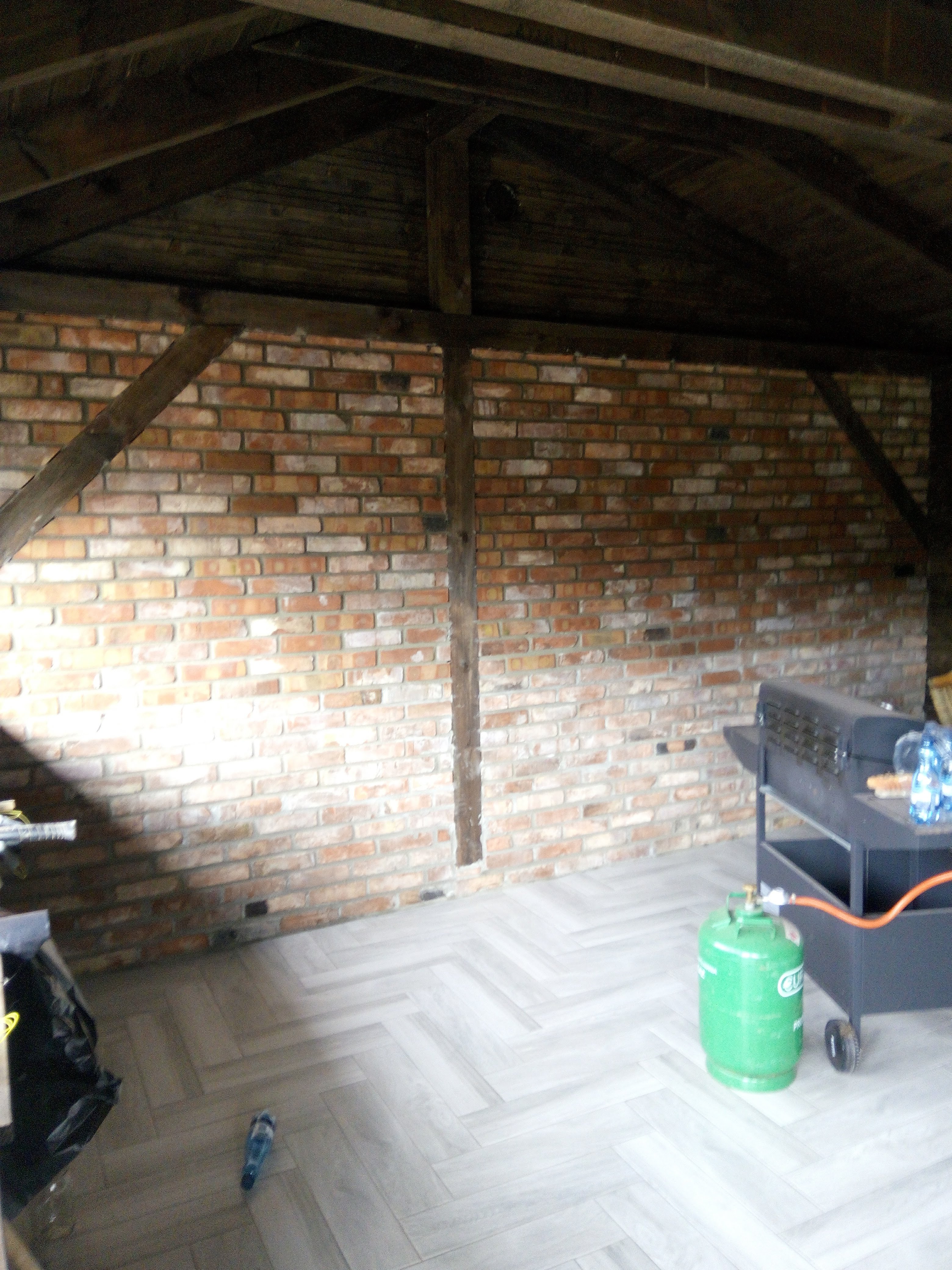 Wnętrze z ceglaną ścianą, drewnianą konstrukcją stropu i podłogą ułożoną w jodełkę. Widoczny grill na kółkach z butlą gazową. Świeżo wykończone pomieszczenie.