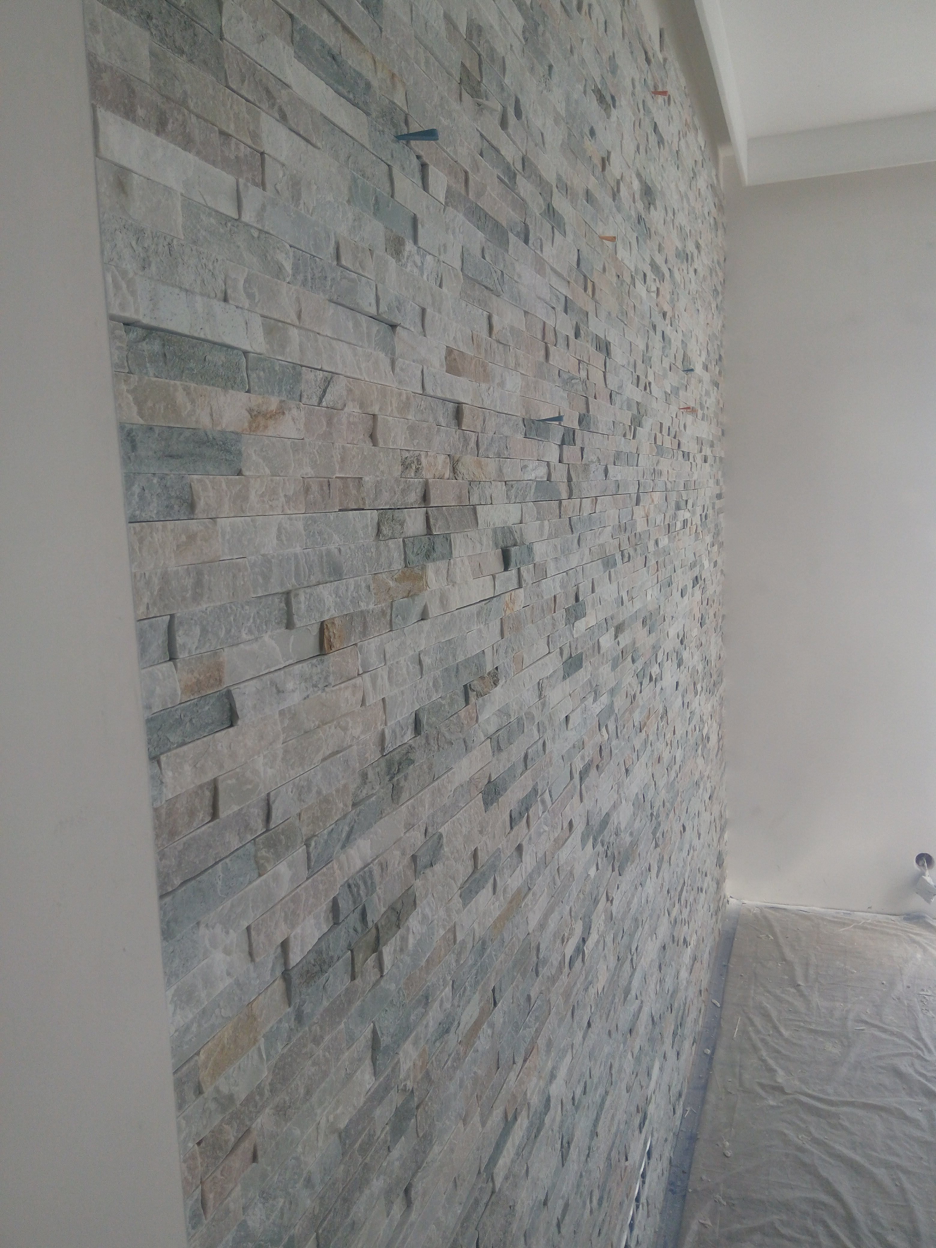 Ściana wyłożona dekoracyjnym kamieniem w odcieniach szarości, beżu i błękitu, z widocznymi plastikowymi klinami montażowymi, zabezpieczona podłoga folią malarską.