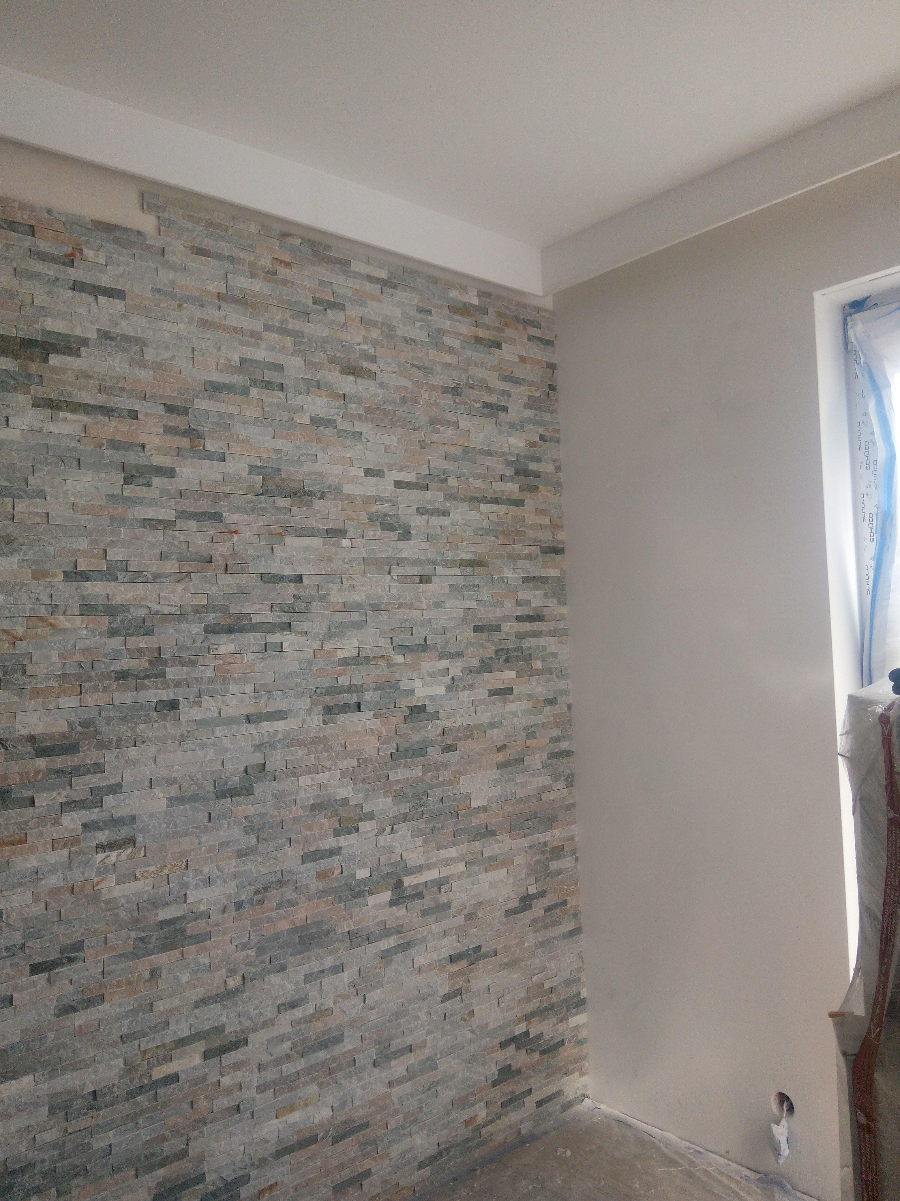 Ściana wykończona dekoracyjną okładziną z kamienia w odcieniach szarości i beżu, częściowo ułożona, z widocznym fragmentem surowej ściany i zakrytym oknem.