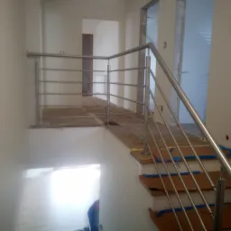 Metalowa balustrada schodowa z poziomymi prętami w jasnym wnętrzu w trakcie montażu, schody zabezpieczone niebieską folią.