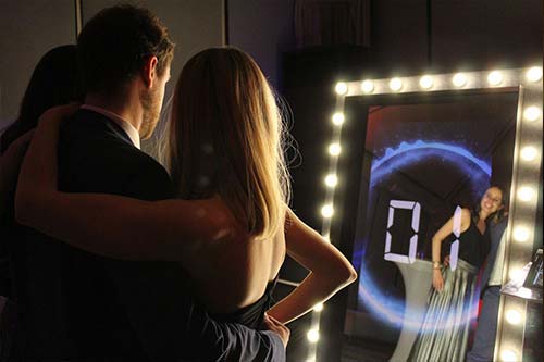 Para obejmująca się przed lustrem z oświetleniem LED, w którym odbija się uśmiechnięta kobieta pozująca do zdjęcia z odliczaniem na ekranie.