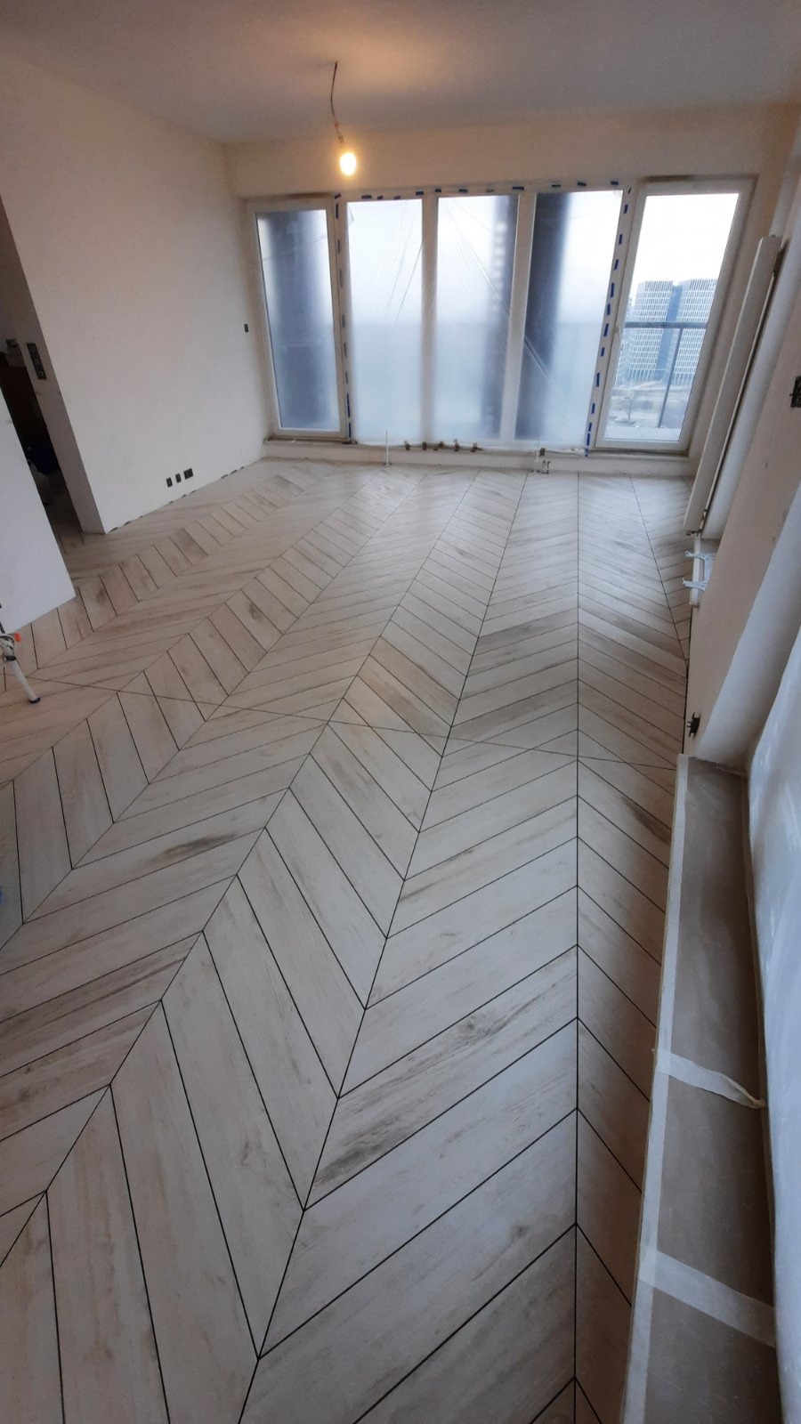 Ułożona w jodełkę podłoga z płytek imitujących drewno w jasnym odcieniu, w pokoju z oknami zabezpieczonymi folią malarską.