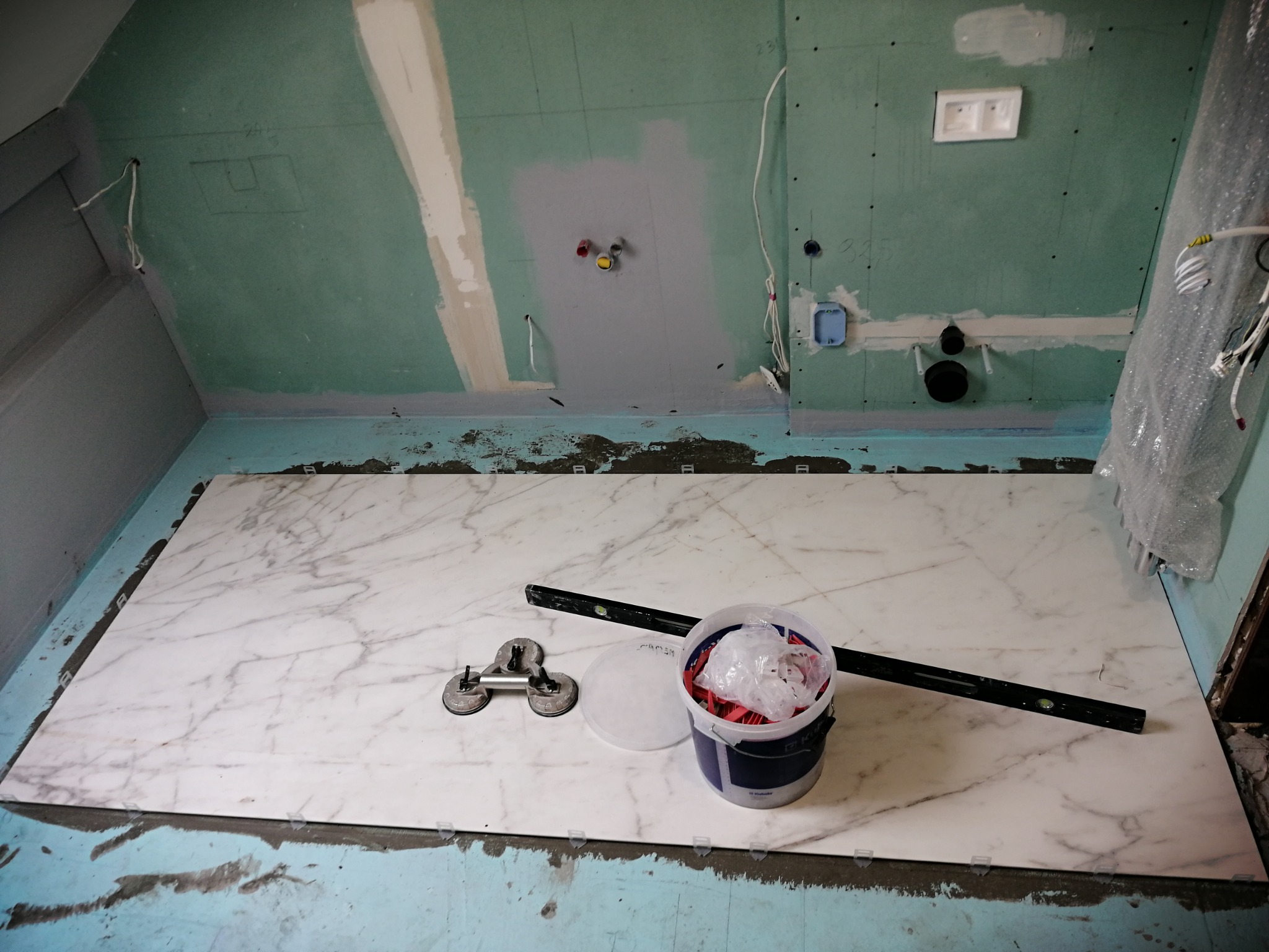 Montaż płyty imitującej marmur na podłodze w łazience, widoczne przyssawki do przenoszenia płyt, poziomica i wiadro z krzyżykami dystansowymi, ściany przygotowane pod płytki.