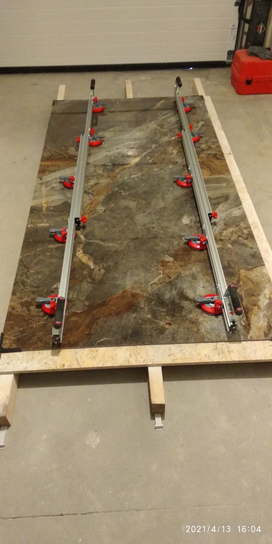 Duża płyta imitująca marmur, zabezpieczona drewnianą ramą i dwiema szynami z przyssawkami, przygotowana do cięcia.