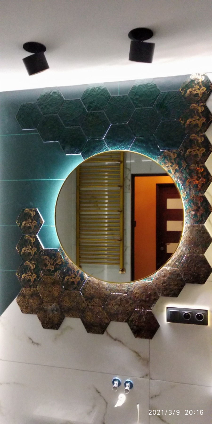 Okrągłe lustro z podświetleniem LED, otoczone dekoracyjnymi, zielono-złotymi heksagonalnymi płytkami, odbijające grzejnik drabinkowy i fragment pomarańczowych drzwi w tle, z czarnymi reflektorami...
