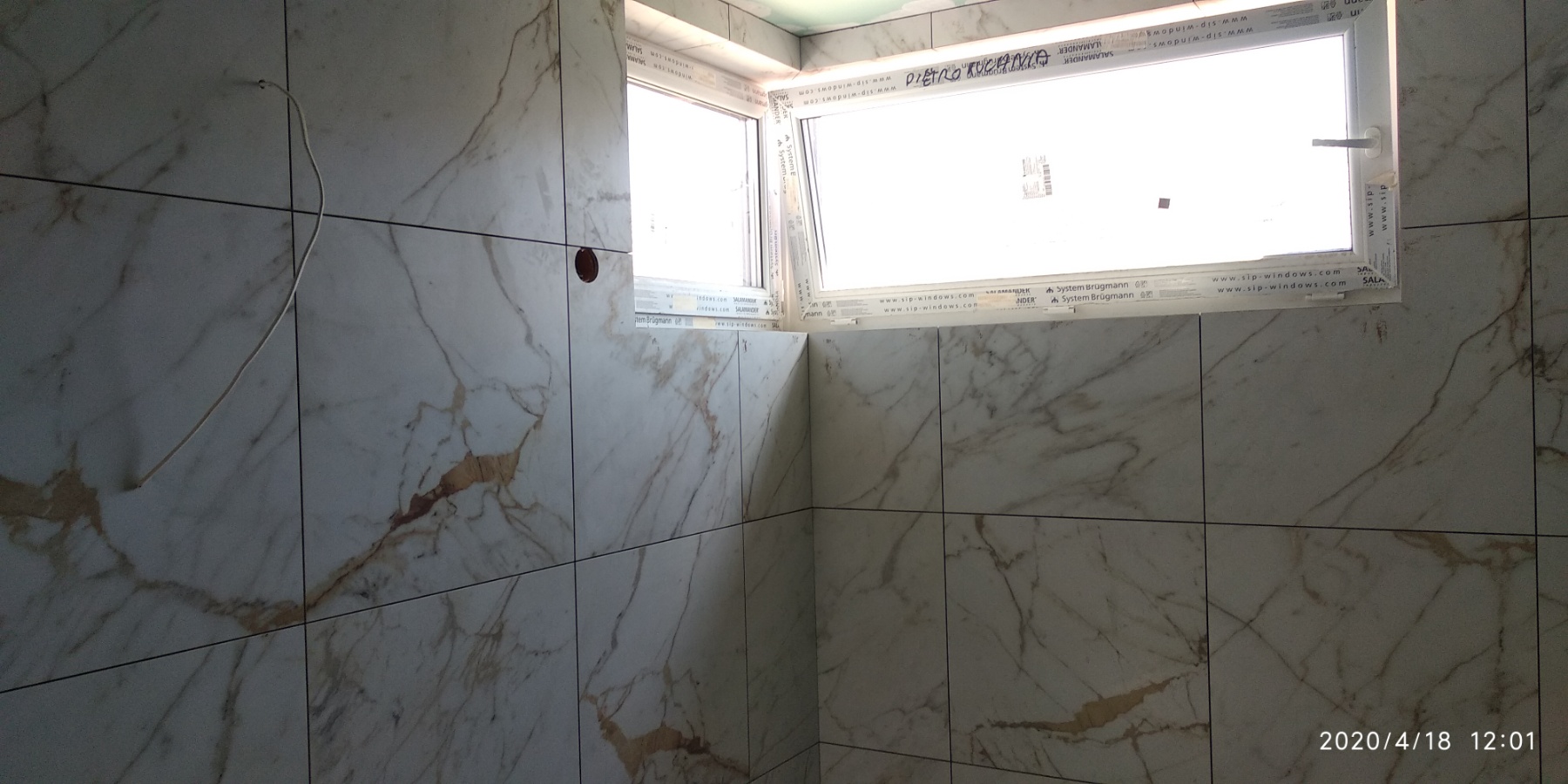 Wnętrze łazienki w trakcie prac wykończeniowych, widoczne okna PCV i ściany wyłożone płytkami imitującymi marmur z widocznym przewodem elektrycznym wystającym ze ściany.