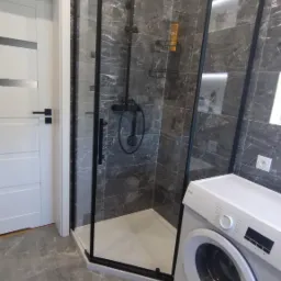 Nowoczesna łazienka z kabiną prysznicową typu walk-in z czarnymi profilami, szare płytki imitujące marmur, biała pralka i mata łazienkowa z wypustkami.