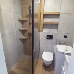 Nowoczesna łazienka z kabiną prysznicową typu walk-in, drewnianymi półkami narożnymi, płytkami imitującymi drewno na podłodze i ścianie w strefie prysznica, oraz pralką w rogu pomieszczenia.