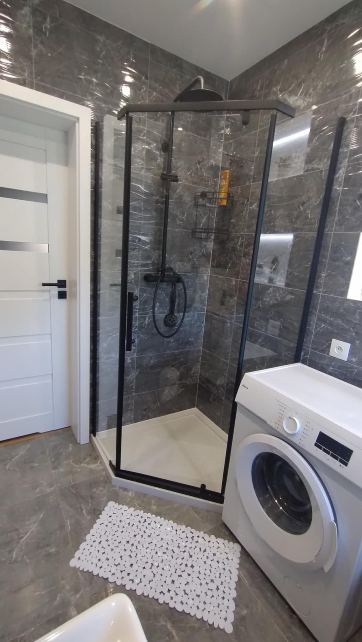 Nowoczesna łazienka z kabiną prysznicową typu walk-in z czarnymi profilami, szare płytki imitujące marmur, biała pralka i mata łazienkowa z wypustkami.