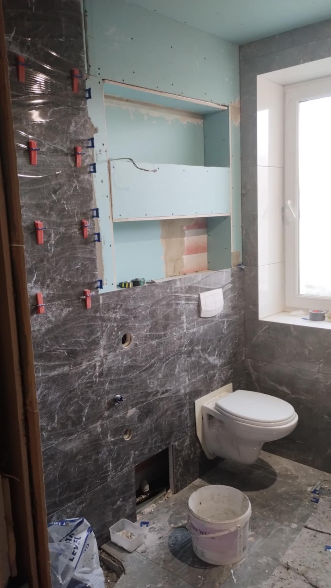 Łazienka w trakcie remontu: szare płytki imitujące marmur, częściowo ułożone, z krzyżykami dystansowymi. W tle widoczna wnęka z płyt karton-gips i podwieszana toaleta.