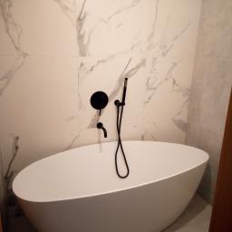 Marcin Szymański USŁUGI REMONTOWO-BUDOWLANE - Wolnostojąca wanna o owalnym kształcie z czarną armaturą na tle ściany wyłożonej płytkami imitującymi marmur.